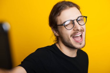 Gözlüklü ve sakallı yakışıklı beyaz adam sarı arka planda kendi portresini yaparken telefon kullanıyor.