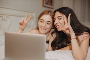 İki neşeli Kafkasyalı kız kardeş yatakta yatıp dizüstü bilgisayar kullanarak biriyle konuşuyorlar.