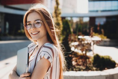 Genç kızıl saçlı beyaz kadın dizüstü bilgisayar tutuyor ve kameraya gözlükleriyle bakıp gülümsüyor.