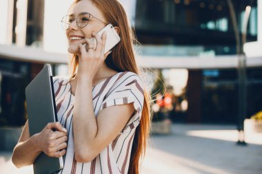 Muhteşem kızıl saçlı beyaz iş kadını elinde dizüstü bilgisayarla telefonda konuşuyor.