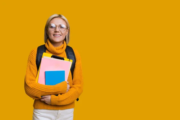 Glada kaukasiska student med blont hår poserar med några böcker och väska på en gul vägg med freespace — Stockfoto