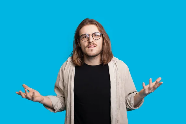 Jovem caucasiano com barba e cabelos longos posando indecisivamente em um fundo azul enquanto olha através de óculos — Fotografia de Stock