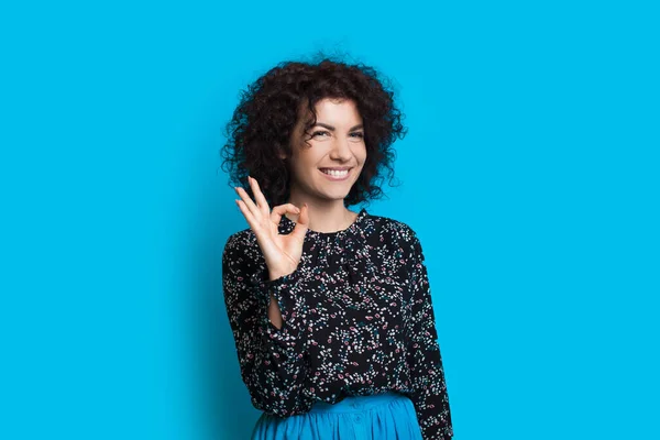 Lockigt hår kaukasiska dam gestikulerar okej tecken medan leende och poserar på en blå bakgrund — Stockfoto
