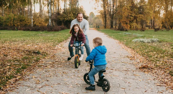 Onlar parkta bisiklete binerken beyaz bir baba çocuklarıyla oynuyor. — Stok fotoğraf