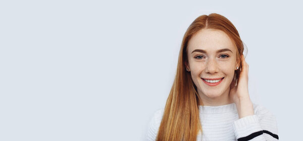 Charming red haired girl with freckles is touching her hair while looking at camera and smiling on white wall with free space