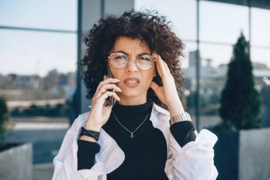 Telefonda konuşurken kıvırcık saçlı ve gözlüklü düşünceli beyaz girişimci poz veriyor.