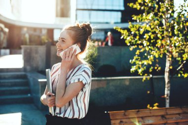 Çekici kızıl beyaz girişimci biriyle telefonda konuşurken dizüstü bilgisayarını tutuyor.