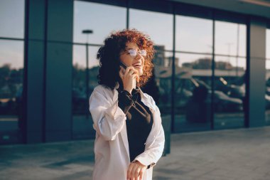 Kıvırcık saçlı ve gözlüklü çekici beyaz kadın binanın önünde poz verirken telefonla konuşuyor.