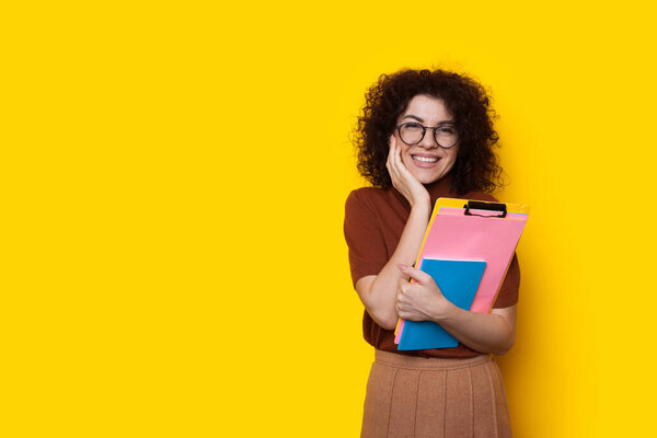 Lovely caucasian student with curly hair and eyeglasses is posing on a yellow wall with free space while holding some books