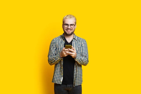 Louro caucasiano homem está conversando com alguém usando um telefone enquanto vestindo óculos em uma parede amarela — Fotografia de Stock