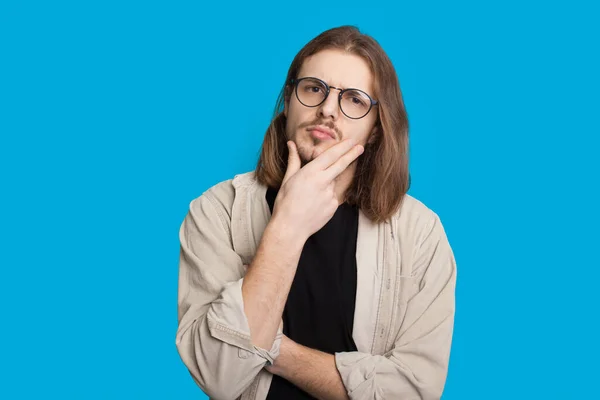 Homem caucasiano cuidadoso com cabelos longos e barba está olhando através de óculos e tocando seu queixo enquanto posando em uma parede azul — Fotografia de Stock