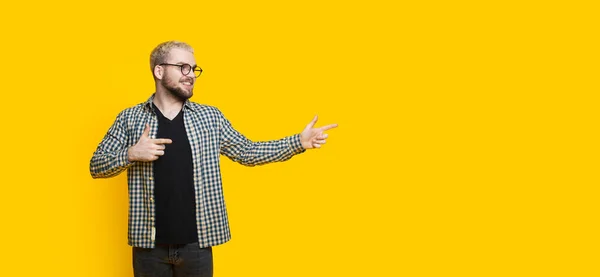 Homem caucasiano loiro com barba está apontando para o espaço livre amarelo perto dele enquanto olha através de óculos — Fotografia de Stock