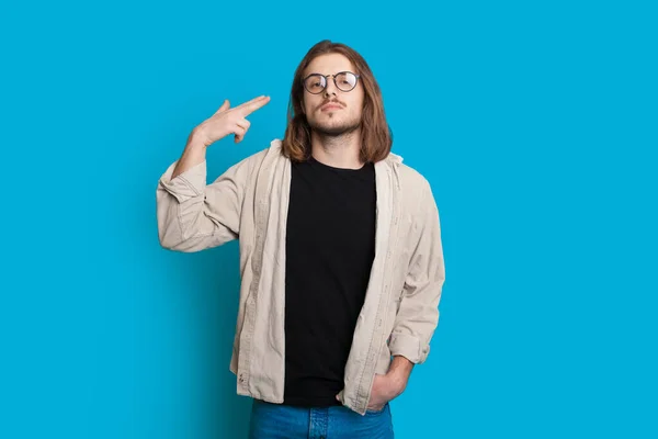 Homem caucasiano com cabelos longos e barba está gesticulando o sinal da pistola apontando para sua cabeça enquanto posando em um fundo azul — Fotografia de Stock