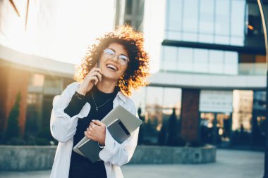 Neşeli beyaz kadın telefonda konuşurken dizüstü bilgisayarıyla gülümsüyor ve poz veriyor.