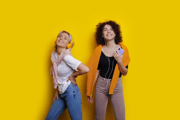 Fröhliche kaukasische Schwestern mit hübschen lockigen Haaren hören Musik über Kopfhörer, während sie auf gelbem Hintergrund posieren — Stockfoto