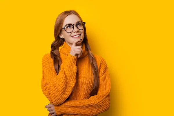 Gânditor ghimbir caucazian doamnă este atinge bărbia ei în timp ce se uită prin ochelari și pozând pe un fundal galben într-un pulover frumos — Fotografie, imagine de stoc