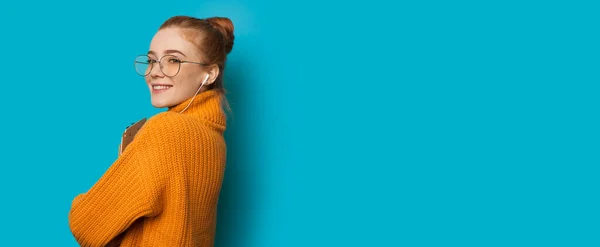 Schöne kaukasische Mädchen mit roten Haaren und Sommersprossen hält ihren Laptop und verwendet Kopfhörer auf einer blauen Wand mit freiem Platz — Stockfoto