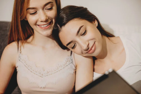 Dos hermanas caucásicas están teniendo alegría juntas mientras sonríen y usan una tableta en el sofá — Foto de Stock