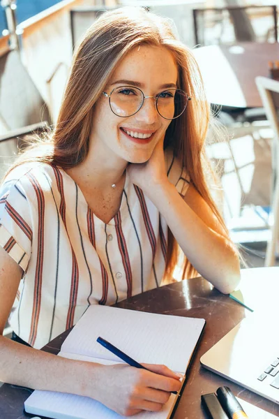 Απολαυστική μαθήτρια με κόκκινα μαλλιά και φακίδες κρατώντας σημειώσεις φορώντας γυαλιά και χρησιμοποιώντας φορητό υπολογιστή σε καφετέρια — Φωτογραφία Αρχείου