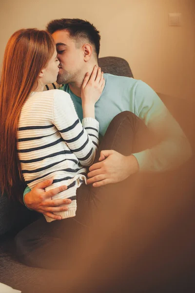 Adorável casal caucasiano beijando uns aos outros enquanto sentado no sofá e abraçando a luz do sol — Fotografia de Stock