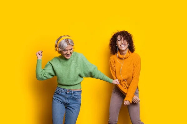 Zwei schöne lockige kaukasische Frauen tanzen auf gelbem Hintergrund, während sie Musik aus Kopfhörern hören — Stockfoto