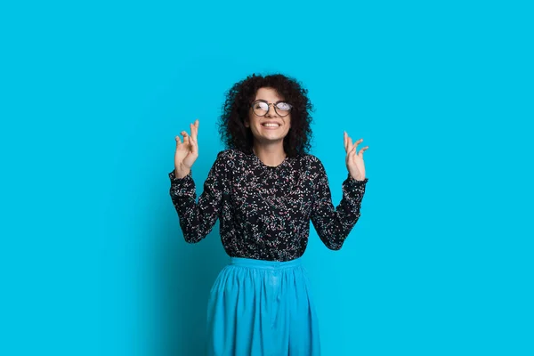 Heerlijk krullend behaarde blanke vrouw met bril wijst omhoog met beide handen terwijl poseren op een blauwe achtergrond — Stockfoto