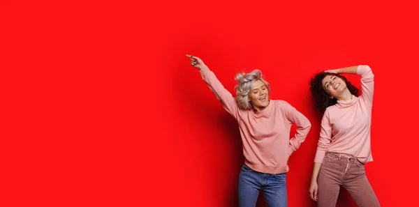 Duas senhoras caucasianas encaracoladas alegres apontando e posando em um fundo vermelho com espaço livre — Fotografia de Stock