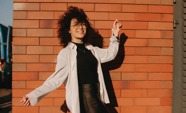 Happy caucasian lady with black curly hair is wearing eyeglasses is posing on a stone wall outside