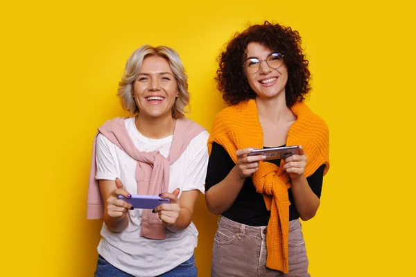Кучеряве волосся біла жінка з окулярами позує в її блондинку сестру, граючи в деякі мобільні ігри на жовтому тлі — стокове фото