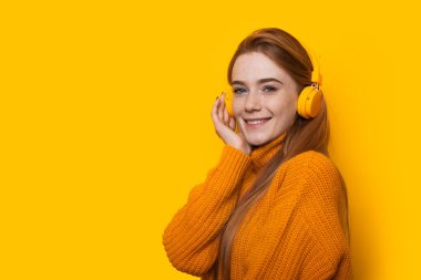 Güzel, kızıl saçlı, çilli bir kız kulaklıkla müzik dinlerken sarı duvarda poz veriyor.