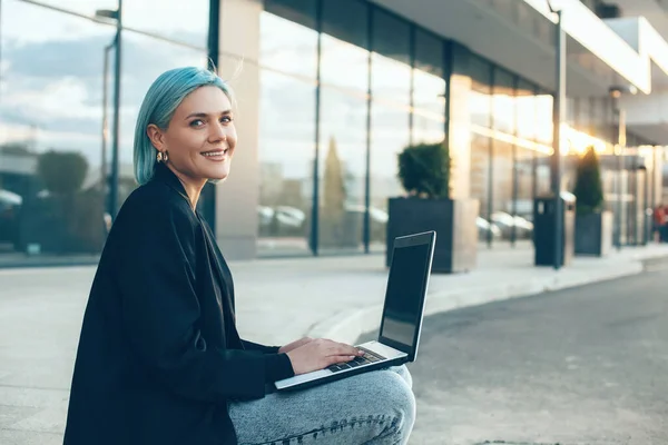 Mavi saçlı beyaz iş kadını büyük bir binanın önünde poz verirken bilgisayarının başında çalışıyor. — Stok fotoğraf