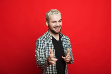 Sarışın ve sakallı yakışıklı beyaz adam kırmızı arka planda kamerayı işaret ediyor.