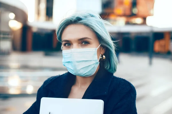 Caucasian entrepreneur with blue hair is wearing a protective mask while posing outside with computer — Stock Photo, Image