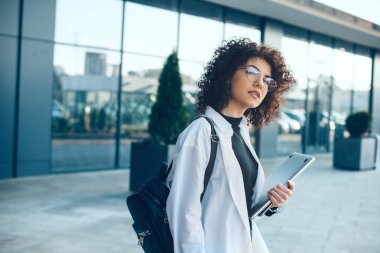 Gözlüklü, kıvırcık saçlı, ciddi bir beyaz öğrenci elinde bir laptop ve çanta tutarken poz veriyor.