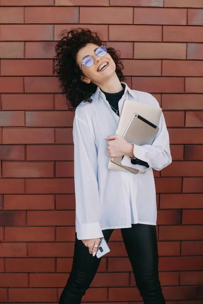 Γλυκιά καυκάσια επιχειρηματίας με σγουρά μαλλιά ποζάρει ευτυχισμένη σε έναν πέτρινο τοίχο ενώ κρατάει τον υπολογιστή της. — Φωτογραφία Αρχείου