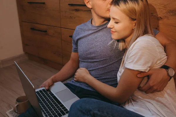 Ljuvligt blond kvinna omfamnar sin älskare medan hon sitter i köket på golvet och använder en bärbar dator — Stockfoto