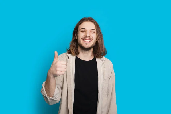 Bonito homem caucasiano com cabelos longos e barba está gesticulando o sinal semelhante em um fundo azul — Fotografia de Stock
