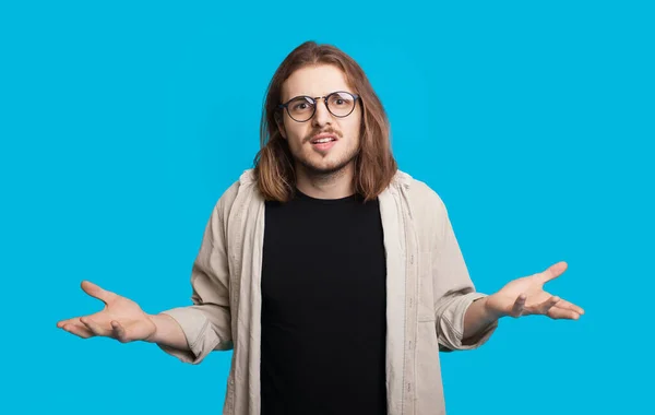 Homem caucasiano com cabelos longos e barba está olhando para a câmera através de óculos e gesticulando uma pergunta em uma parede azul — Fotografia de Stock