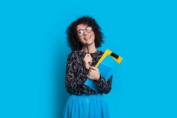 Charmante lockige Studentin hält ein paar Bücher in der Hand, während sie in einem Kleid auf blauer Wand mit Leerraum posiert — Stockfoto