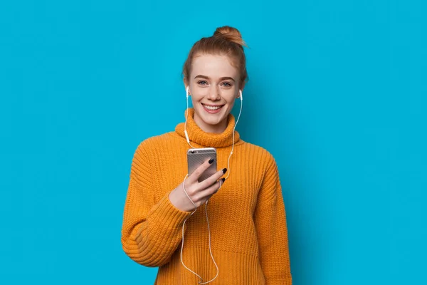 Ginger lady con pecas sosteniendo un teléfono y escuchando música mientras sonríe sobre fondo azul — Foto de Stock