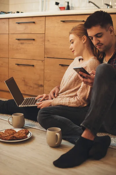 Kedves kaukázusi pár ül a konyhában a padlón böngészve egy laptop és telefon, miközben a reggeli — Stock Fotó