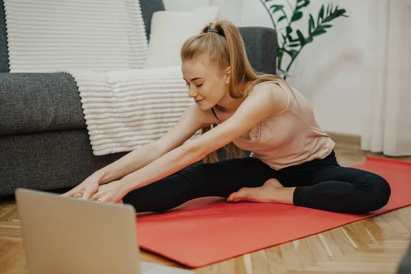 Chica caucásica pelirroja con pecas estirándose en una alfombra roja en el suelo mientras hace yoga cerca de una computadora portátil —  Fotos de Stock