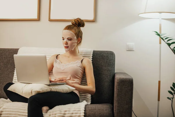 Mujer caucásica con el pelo rojo lleva una máscara de limpieza en la cara mientras se sienta en el sofá y el uso de un ordenador portátil — Foto de Stock