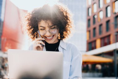 Kıvırcık saçlı neşeli beyaz girişimci bilgisayarında çalışırken telefon görüşmesi yapıyor.