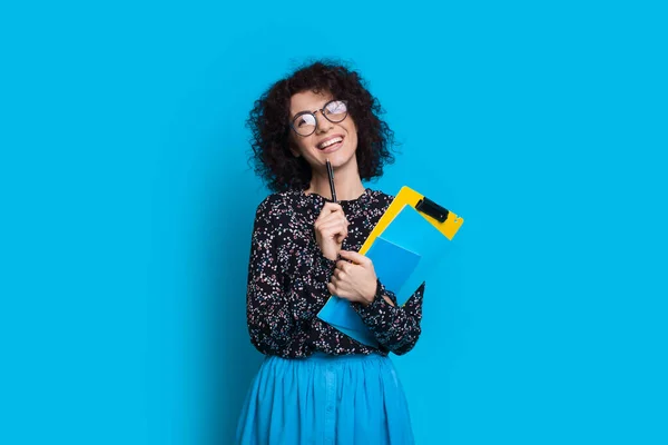 Charming curly haired student holding some books while posing in a dress on blue wall with blank space — Stock Photo, Image