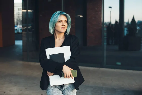 Affascinante donna d'affari caucasica con i capelli blu sta guardando lontano mentre tiene un computer portatile e un telefono davanti a un edificio — Foto Stock