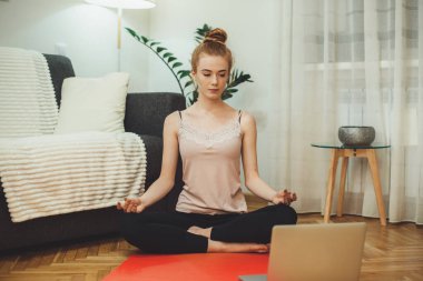 Kızıl saçlı, çilli bir kız yerde dizüstü bilgisayar kullanarak yoga dersleri alıyor.