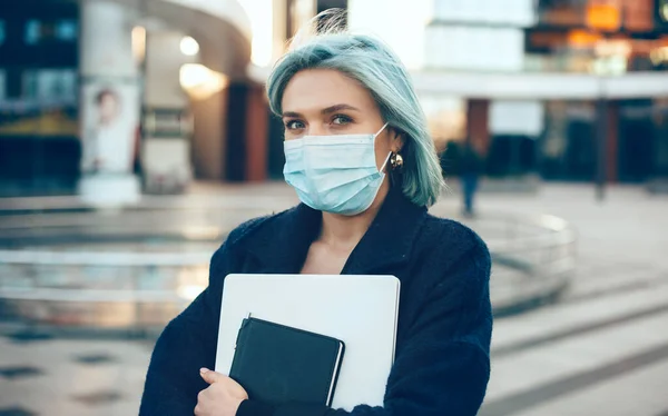 Blue haired caucasian entrepreneur holding a laptop while wearing a special white mask and looking at camera — Stok Foto