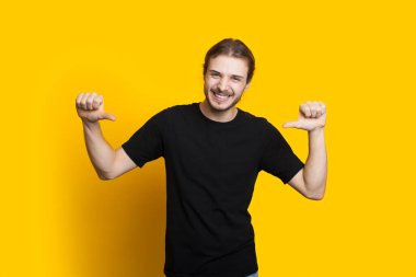 Sakallı, uzun saçlı, kendine güvenen beyaz bir adam sarı arka planda poz verirken onu işaret ediyor.