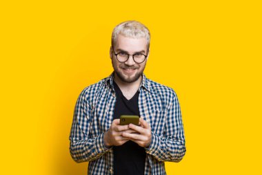 Sarı saçlı ve gözlüklü beyaz çocuk kameraya bakıyor. Sarı duvardaki biriyle konuşuyor.